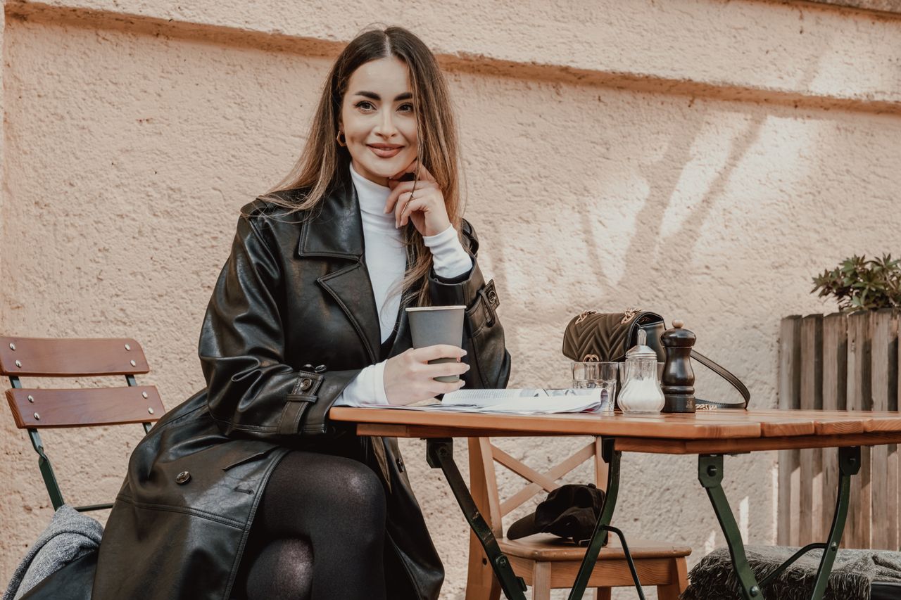 services-01 Woman in a black coat smiling while enjoying coffee outdoors at a cafe table.