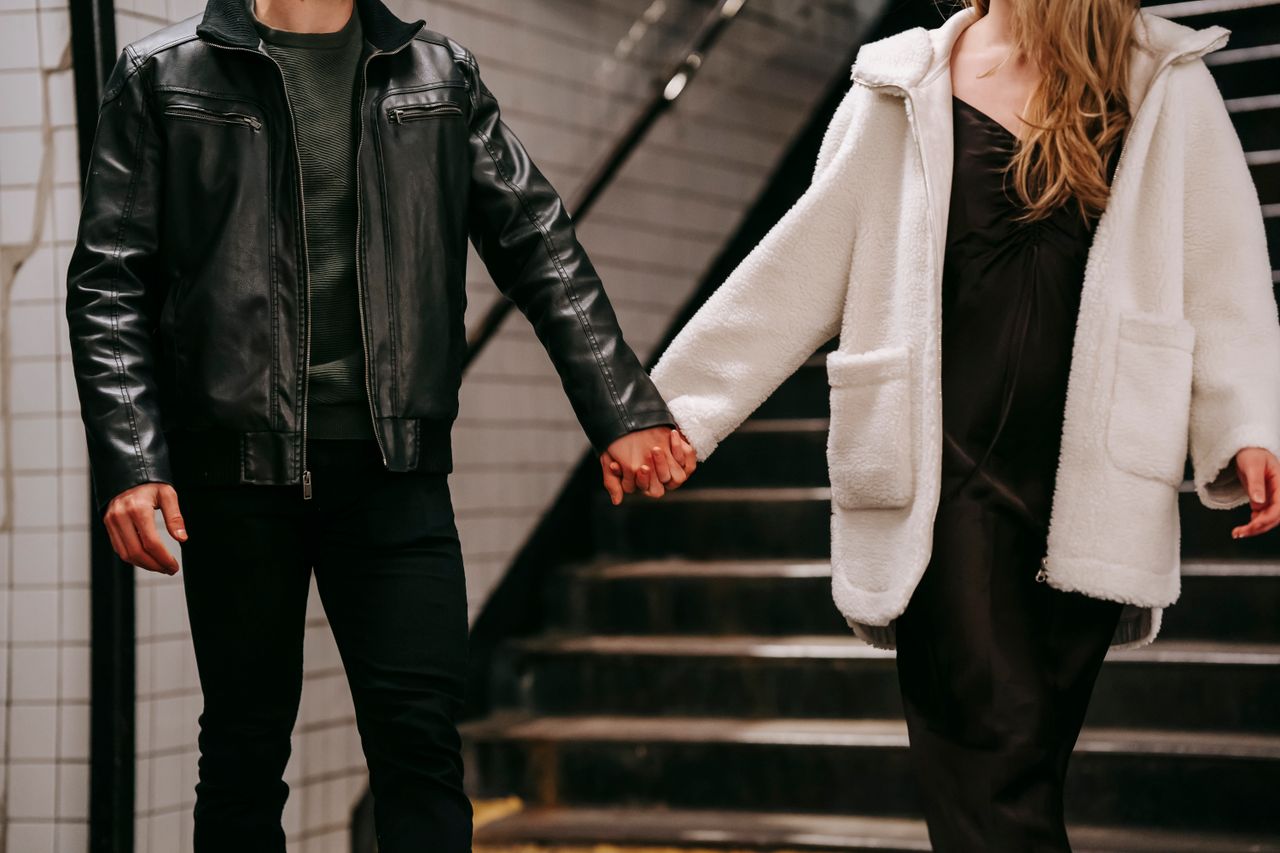 Unrecognizable boyfriend and girlfriend wearing warm clothes holding hands while walking in subway station with metal stairway during romantic date