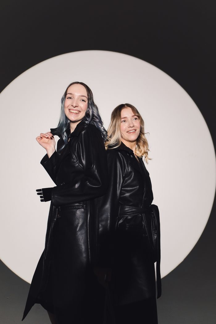 ours-journey Two stylish women posing back to back in trendy black leather coats against a spotlight.